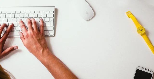 Hands typing on a keyboard, with a mouse, watch, and phone nearby.