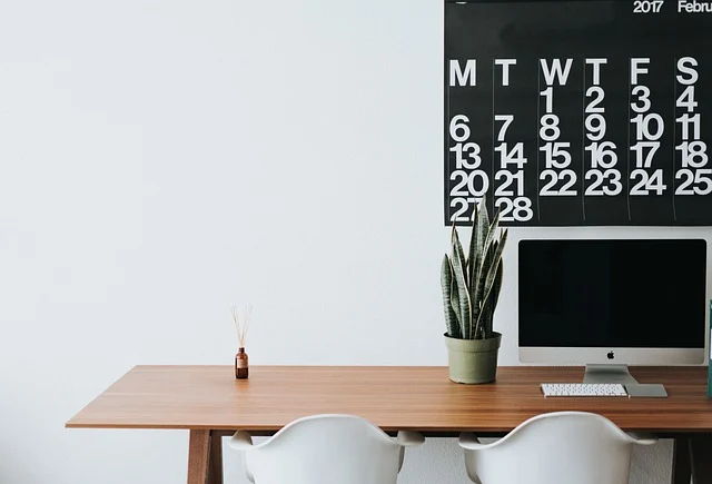 A clean desk with a computer, plant, and calendar on the wall.