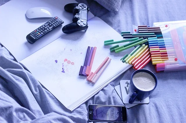 A game controller, remote, mouse, phone, markers, and coffee on a bed.