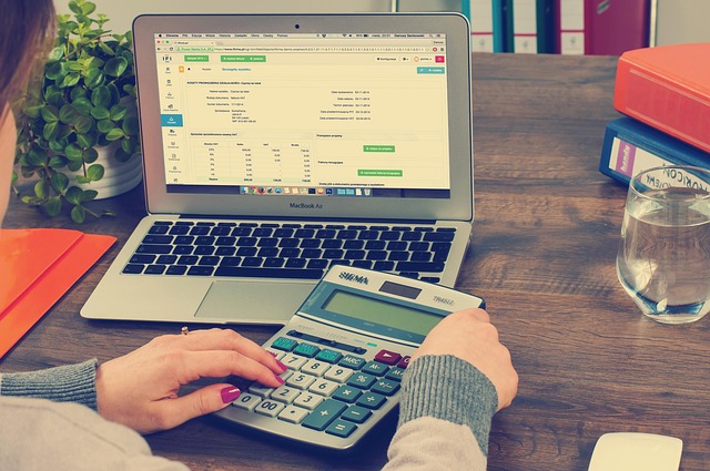 A person using a calculator next to a laptop displaying financial data