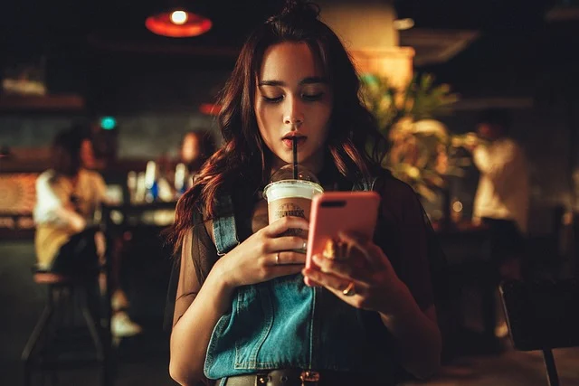 A woman drinking coffee and looking at her phone in a dimly lit cafe.