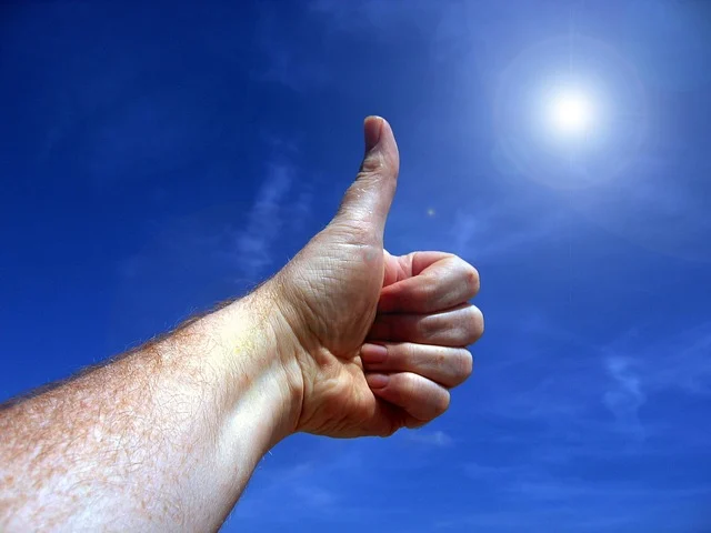 A hand giving a thumbs-up gesture against a blue sky with the sun.