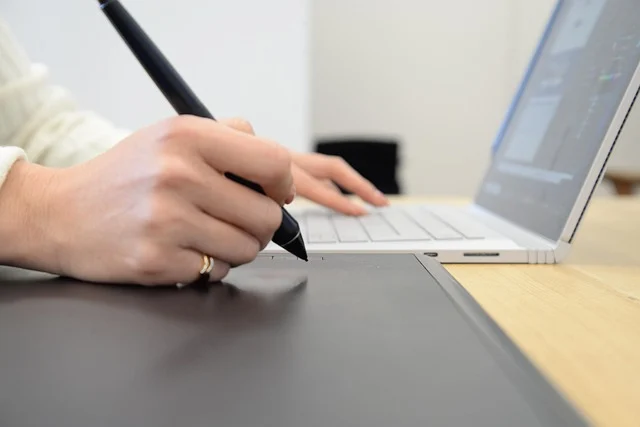 A person's hands using a stylus on a tablet and typing on a laptop.