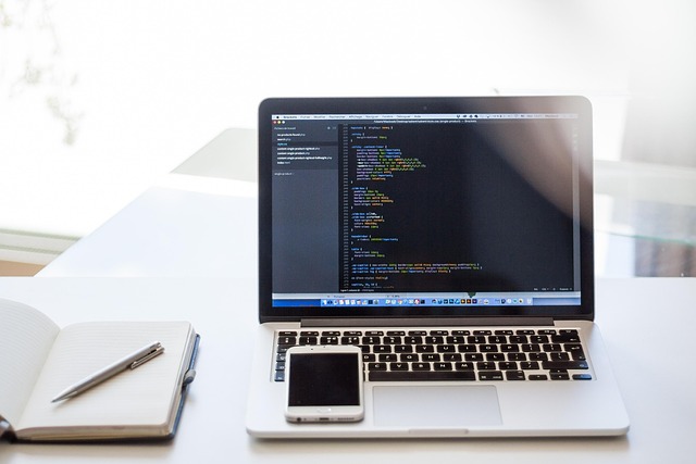 A laptop displaying code next to a smartphone and notebook