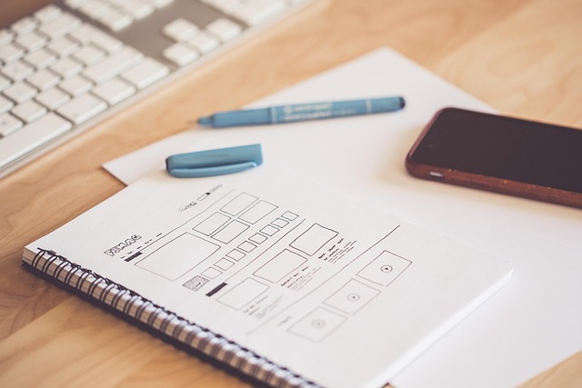 A notebook with app wireframe sketches, a smartphone, and a keyboard