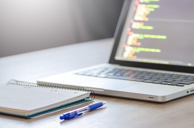 A laptop displaying code next to a notebook and pen