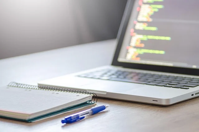 A laptop displaying code next to a notebook and pen