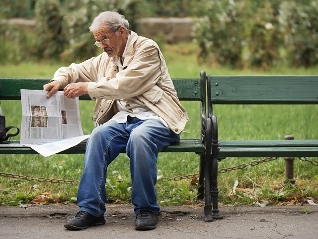 An elderly man sitting on a park bench reading a newspaper