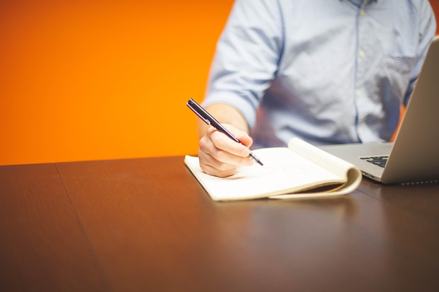 A person writing in a notebook next to a laptop at a desk