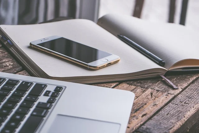 A MacBook, iPhone, notebook, and pen on a wooden table.