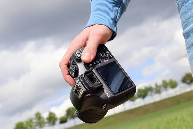 A hand holding a DSLR camera outdoors under a cloudy sky