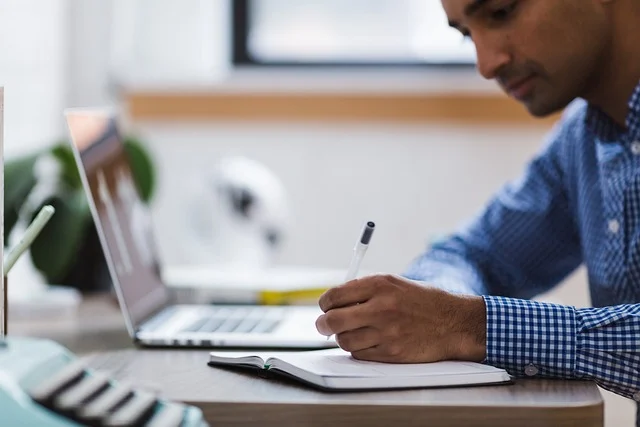A man writing in a notebook with a laptop in the background.