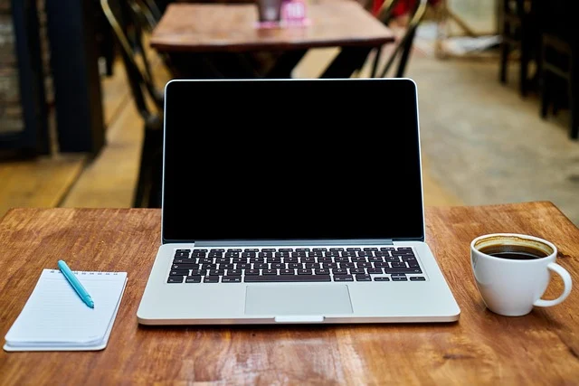 A MacBook Pro on a table with a blank screen, coffee, and a notebook.