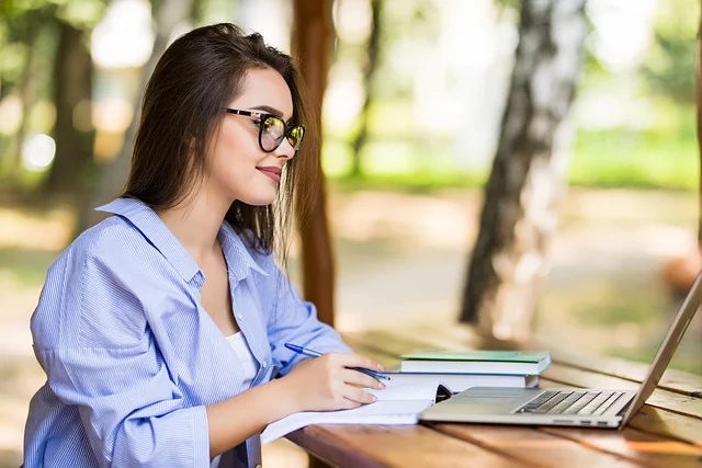 A woman working on a laptop and writing in a notebook outdoors.