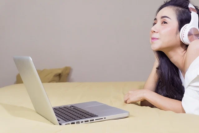 A woman wearing headphones and looking at a laptop on a bed.